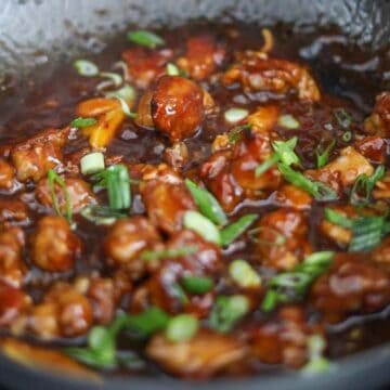 Fried chicken added to sauce in pan, tossed to coat evenly, garnished with green onions and sesame seeds, ready to serve