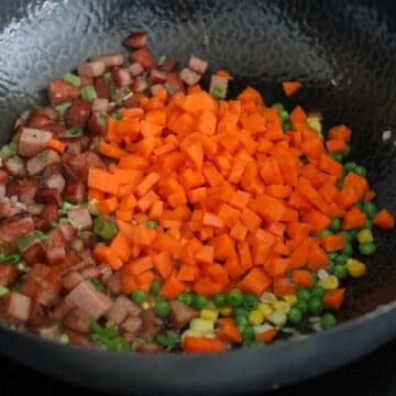 A wok with aromatics and vegetables being stir-fried-green onions, garlic, corn, peas, and carrots-tossed together before being pushed to one side of the pan.