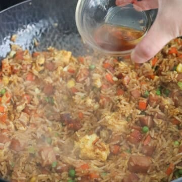 Cold rice added to a wok with soy sauces drizzled around the edges, sizzling as butter melts in. The rice is being tossed with seasonings until evenly coated and finished with sesame oil and green onions.