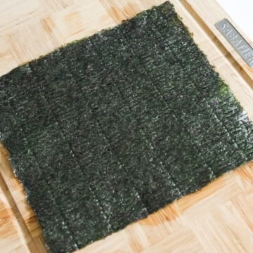 On a clean working surface, lay out a sheet of roasted seaweed rough side up. Evenly sprinkle salt, garlic powder, onion powder and sesame seeds on top of the seaweed.
