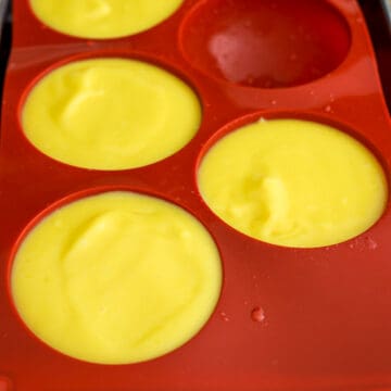 Mango mixture being poured into greased bowls or silicone molds, with air bubbles tapped out and surface smoothed.
