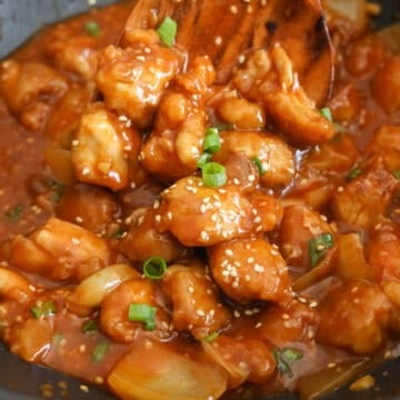 Crispy fried chicken being tossed in thickened sauce in a pan until fully coated and glossy, finished with green onions and sesame seeds.