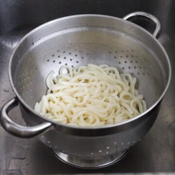 Udon noodles soaking in a large bowl of boiling water until loosened, then drained.