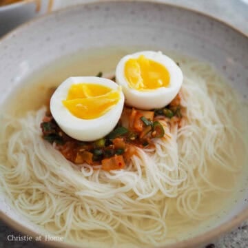Noodles divided into bowls, topped with sauce and halved eggs, with hot broth poured over before serving.