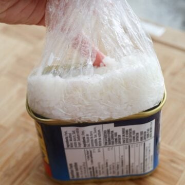 Cover the rice with the plastic wrap. Then using your fingers, pack the rice into base of can until top is flat. Be careful not to cut yourself along the sharp edges of the can, feel free to use a utensil to help you with this step. Remove the block of white rice from can by pulling the cling film out and unwrap it on a cutting board.