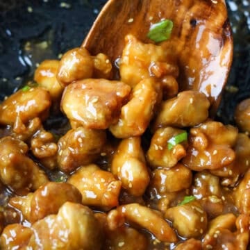 Thickened orange sauce bubbling in a pan as crispy chicken is added and tossed to coat, finished with orange zest, sesame seeds, and green onions.