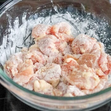 Diced chicken thighs in a bowl being seasoned with salt and garlic powder, then coated evenly with cornstarch.