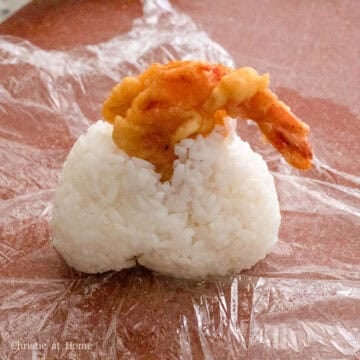 Insert the seasoned shrimp tail side up into the hole of the rice ball. The rice will break apart here which is normal, so cover the ball with the plastic wrap and shape it back into a triangle.