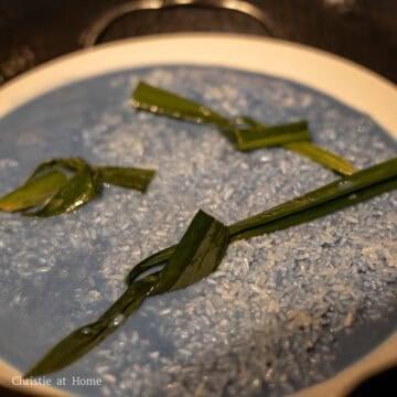 Next pour the blue rice and the liquids onto a large shallow bowl. Then place knotted pandan leaves into the rice. Fill a large wok or steamer pot halfway with water and place a wire rack or bamboo steamer inside. Bring to a boil on medium-high heat and then reduce to medium heat. Carefully place the plate of blue rice on top of the wire rack or bamboo steamer.