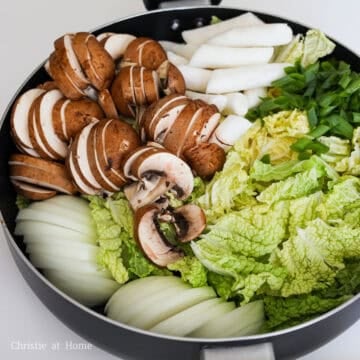 Place prepared onion, green onion, cabbage, mushrooms, spam, tofu, maple baked beans, kimchi and rice cakes into large pan that is about 3 inches deep.